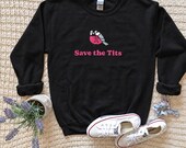 Photo 7 of 20: A black crewneck sweatshirt with a printed bird graphic and the text Save the Tits is displayed flat with white sneakers, a potted plant, and a scrunchie on a knitted throw.