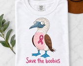 Photo 9 of 17: A folded white t-shirt printed with a cartoon blue-footed booby bird graphic, pink ribbon, and the text Save the boobies.