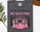 Photo 5 of 17: A folded charcoal grey t-shirt showing the In October We Wear Pink witch artwork, set beside green leaves and a brown hat.