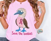 Photo 1 of 17: A woman wearing a pink t-shirt featuring a cartoon blue-footed booby bird with a pink ribbon and the text Save the boobies.