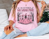 Photo 8 of 17: A person sits on a bed wearing a light pink t-shirt with a witch graphic and the text In October We Wear Pink, paired with distressed jeans.