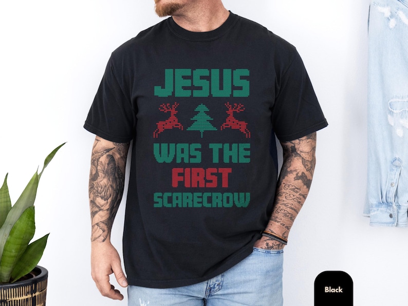 Funny christian tee with a black shirt and retro holiday graphic reading Jesus was the first scarecrow, styled like a cozy ugly sweater style design with festive reindeer and tree details, perfect christian gift shirt or gag gift shirt for Christmas laughs