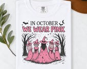 Photo 7 of 17: A folded white t-shirt featuring the In October We Wear Pink witch graphic, arranged next to green foliage and a brown hat.