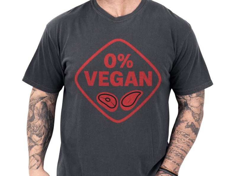 Charcoal vintage soft cotton tee with bold distressed red 0% VEGAN diamond graphic and steak icons, shown on a casually tattooed model — a funny BBQ shirt and grilling tee for backyard cookouts, ideal meat lover gift, grillmaster shirt and casual BBQ tee or gift for dad