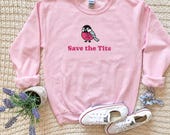 Photo 4 of 20: A light pink crewneck sweatshirt with a bird graphic and the text Save the Tits is arranged flat alongside white sneakers, a small potted plant, and a scrunchie on a textured blanket.