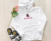 Photo 6 of 20: A white hoodie with a graphic of a bird wearing a pink ribbon and the text Save the Tits is arranged on a wooden floor with black and white sneakers and a stem of pink roses.