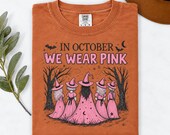 Photo 6 of 17: A folded rust orange t-shirt displaying the In October We Wear Pink witch design, alongside a leafy branch and a brown hat on a white background.
