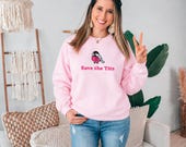 Photo 2 of 20: A smiling woman wears a light pink sweatshirt with a bird graphic and the text Save the Tits while posing in a decorated room.