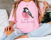 Photo 2 of 17: A person sitting on a bed wearing a pink graphic t-shirt featuring a bird and a pink ribbon.