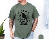 Photo 5 of 8: Moss green unisex crow pun tshirt on a relaxed model, soft vintage wash feel, bold raven graphic shirt with a crow perched on a skull and bare branches and F-CAW-F text, a gothic crow shirt / skull bird tee with dark humor and edgy humor vibes, ideal gifts for men