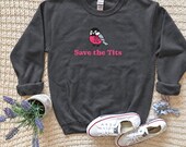 Photo 9 of 20: A dark heather gray crewneck sweatshirt with a bird graphic and the text Save the Tits is arranged flat with white sneakers, a small plant, and a patterned scrunchie on a textured fabric surface.
