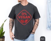 Photo 2 of 8: Charcoal casual tee featuring a bold red 0% VEGAN badge with steak icons, worn by a tattooed model for relaxed backyard grilling — a funny BBQ shirt and grillmaster shirt pick, ideal as a meat lover gift or backyard BBQ tee and a playful gift for dad who loves to smoke and grill