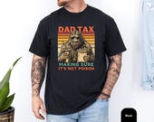 Photo 3 of 8: Man wearing a vintage-washed pepper tee with a retro sunset Sasquatch holding fries and a drink and a cheeky "Dad Tax" joke, playful dad joke tee and sasquatch shirt perfect as a funny dad gift tee or casual weekend camping dad shirt