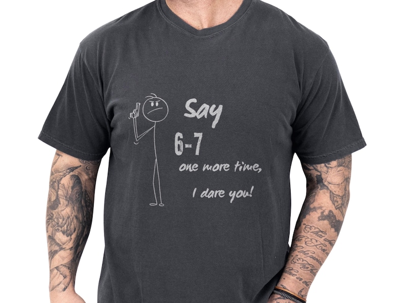 Charcoal minimalist stick figure tee showing a hand‑drawn stick man and cheeky script saying Say 6‑7 one more time I dare you, worn by a casually tattooed model — playful, youthful casual tee perfect as a teacher gift tee, parent gag gift shirt, or humorous gift for teacher appreciation