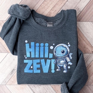 May include: A dark gray sweatshirt with the text "Hiiii, ZEV!" in blue. The sweatshirt features a cartoon illustration of a blue fish-like creature in a spacesuit. The design is centered on the front of the garment.