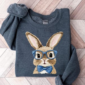 May include: A dark grey sweatshirt with an embroidered rabbit wearing blue spectacles and a blue bow tie. The rabbit has tan fur and long ears. The sweatshirt is folded on a light wood grain surface.