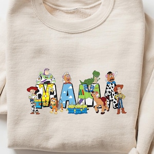May include: A beige sweatshirt featuring the word "MAMA" in colorful block letters, each letter decorated with characters from the animated film Toy Story. The sweatshirt has long sleeves and a crew neck.