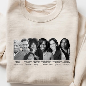 May include: Cream-colored sweatshirt featuring a black and white graphic of six women with the text "Harriet, Ida, Rosa, Ruby, Michelle, Kamala, Retana, Could, So" and their signatures. The sweatshirt is folded.