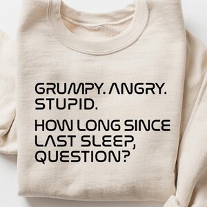 May include: A cream-colored sweatshirt with the text "GRUMPY. ANGRY. STUPID. HOW LONG SINCE LAST SLEEP, QUESTION?" in black, centered on the front. The sleeves are folded up, and the fabric appears soft.