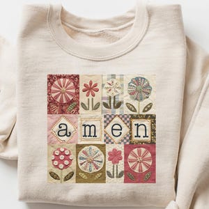 Peut inclure: Sweat-shirt crème avec un motif floral et le mot "amen". Le motif comprend divers motifs floraux dans les tons de rouge, rose et vert. Le sweat-shirt a des manches longues et un col rond.