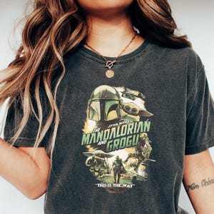 Puede incluir: Camiseta gris oscuro con un dise&ntilde;o gr&aacute;fico de la serie Star Wars, "The Mandalorian and Grogu". El dise&ntilde;o incluye el casco mandaloriano, Grogu y la frase "THIS IS THE WAY". La camiseta se usa con vaqueros azules y zapatillas blancas.