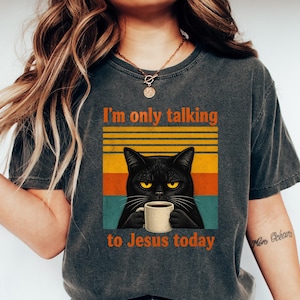 May include: Dark grey t-shirt with the text "I'm only talking to Jesus today." The shirt features a graphic of a black cat holding a coffee mug. The cat is set against a retro sunset design.