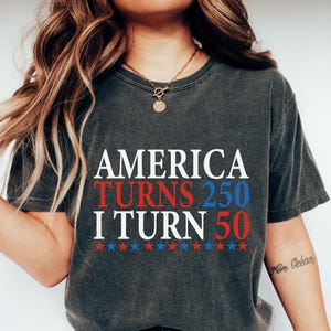 May include: Dark gray t-shirt with the text "AMERICA TURNS 250 I TURN 50" in white, red, and blue. The shirt is paired with blue jeans and white sneakers. The overall theme is patriotic and celebratory.