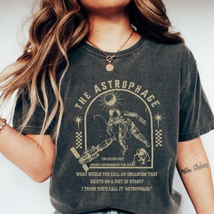 May include: Dark gray t-shirt with a graphic featuring an astronaut, sun, and text: "THE ASTROPHAGE." The design includes the text "THE SILVER DUST, ENERGY DEVOURER OF THE STARS" and a question about organisms that eat stars. The shirt is worn with blue jeans and white shoes.