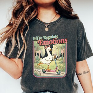 Peut inclure: T-shirt gris foncé avec l'inscription « Off to Regulate Emotions ». Le graphisme représente une oie de dessin animé sur une trottinette rose, tenant une tasse de café. L'oie porte un nœud et se trouve dans un décor de parc.