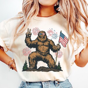 Pu&ograve; includere: Una t-shirt color crema con la grafica di un Bigfoot marrone che indossa occhiali da sole con il motivo della bandiera americana, fa il segno di pace e tiene in mano una bandiera americana. Il design include fuochi d'artificio e pini.