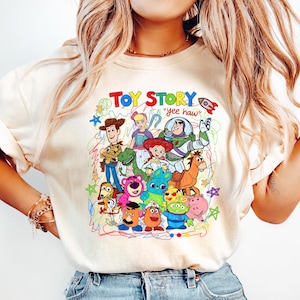 May include: Cream-colored t-shirt featuring a colorful graphic of Toy Story characters, including Woody, Buzz Lightyear, and others. The text "TOY STORY" is at the top, with "yee haw" below. The shirt is paired with light blue jeans.