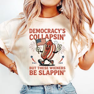 May include: A cream-colored short-sleeve t-shirt featuring a graphic of a cartoon hot dog holding an American flag and making a peace sign. The text reads DEMOCRACY'S COLLAPSIN' BUT THESE WIENERS BE SLAPPIN' in red and blue lettering with star accents.