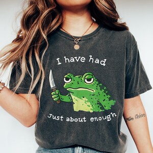 May include: Dark gray t-shirt featuring a green frog holding a knife. The text on the shirt reads "I have had Just about enough". The frog has a grumpy expression. The shirt is paired with blue jeans and white shoes.