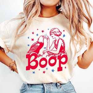 May include: A cream-colored short-sleeve t-shirt featuring a red and blue graphic print. The design depicts a person touching the beak of a bald eagle, surrounded by small stars, with the word Boop printed in large red letters underneath.