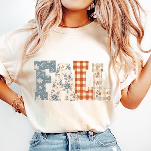 May include: A cream-coloured short-sleeve t-shirt featuring the word FALL printed in large block letters across the chest. Each letter has a distinct pattern: the F is blue floral, the A is blue floral, the L is orange gingham, and the L is plaid.