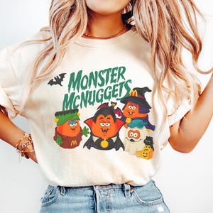 May include: A cream-colored short-sleeve t-shirt featuring a graphic print of four cartoon characters dressed as monsters with the text Monster McNuggets in green. The characters include a Frankenstein, a vampire, a witch, and a ghost.