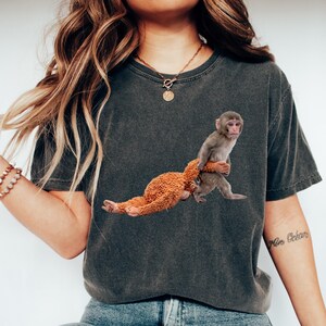 May include: Dark gray t-shirt featuring a graphic of a monkey holding a stuffed animal. The monkey is gray and the stuffed animal is brown. The shirt is worn with blue jeans and white sneakers.