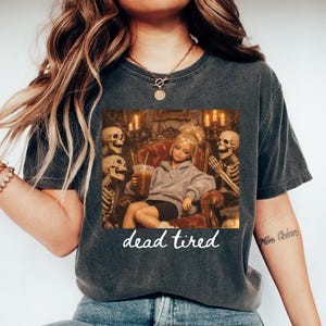 May include: A charcoal gray short-sleeve t-shirt featuring a graphic print of a doll in a hoodie sitting in a chair surrounded by skeletons, holding a beverage. The text "dead tired" is printed in white script below the graphic illustration.