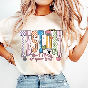 May include: A cream-coloured t-shirt with the words "TEST DAY" in colourful, patterned letters. The shirt also features the text "don't stress do your best!" and includes images of a pencil, apple, and calculator.