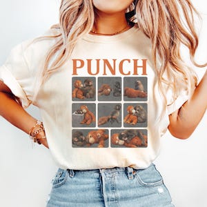 May include: Cream-colored t-shirt with the word "PUNCH" in orange block letters. Below the text are nine small squares, each containing a vintage-style illustration of teddy bears in various poses. The shirt is paired with light blue jeans.