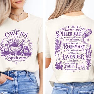 May include: A cream-colored t-shirt featuring purple graphics. The front displays a mortar and pestle with the text Owens Sisters Apothecary Est. 1995. The back features text about spilled salt, rosemary, and lavender with botanical illustrations.