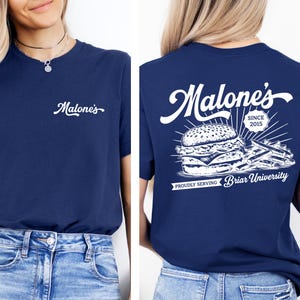 May include: Navy blue t-shirt with the word "Malone's" in white script on the front. The back of the shirt features a burger and fries graphic with the text "Malone's Since 2015" and "Proudly Serving Briar University."