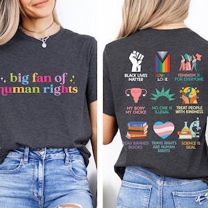 May include: Dark gray t-shirt with the text "big fan of human rights" in colorful letters. The back of the shirt features various social justice slogans and symbols, including "Black Lives Matter" and "Trans Rights are Human Rights."