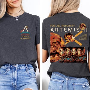 May include: Dark gray t-shirt featuring the Artemis logo on the front and a graphic on the back with the text "FOR ALL HUMANITY ARTEMIS II". The back graphic includes an illustration of astronauts and a spacecraft.