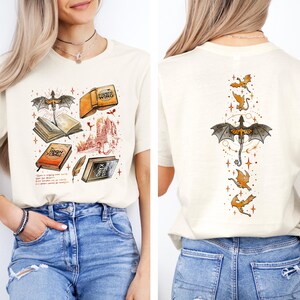 May include: Cream-colored t-shirt featuring a fantasy-themed illustration on the front, including books, a castle, and a dragon. The back of the shirt displays a dragon and several smaller dragons. The design incorporates orange, black, and brown elements.