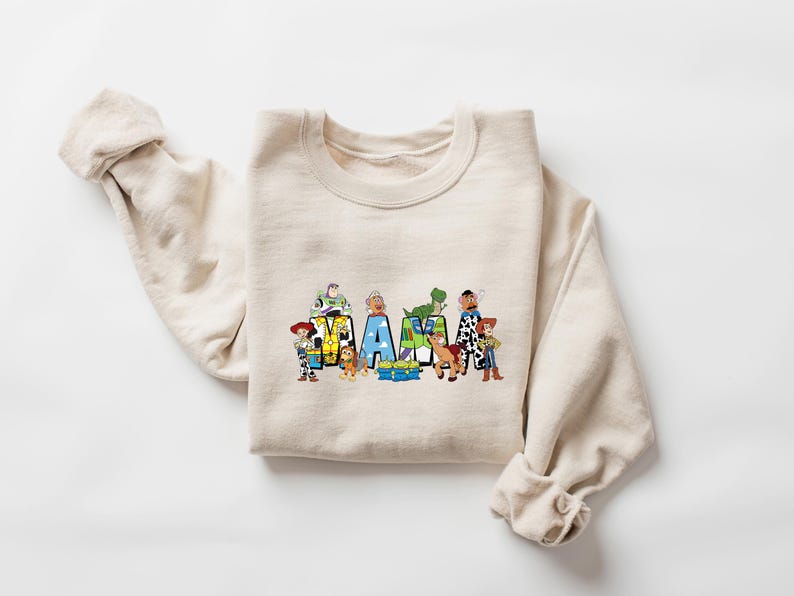 May include: A beige sweatshirt featuring the word "MAMA" in colorful block letters, each letter decorated with characters from the animated film Toy Story. The sweatshirt has long sleeves and a crew neck.