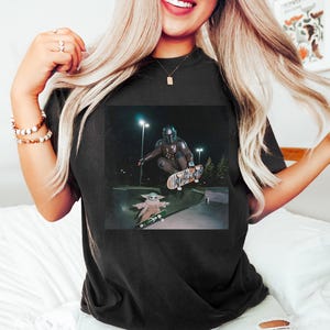 Puede incluir: Camiseta negra con un gr&aacute;fico de The Mandalorian y Grogu patinando en un skatepark por la noche. La imagen muestra a los personajes en el aire sobre monopatines, con Grogu en una tabla m&aacute;s peque&ntilde;a.