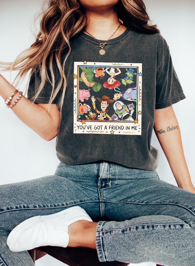 Op de afbeelding: Een antracietgrijs t-shirt met korte mouwen en een vierkante grafische print van diverse geanimeerde speelgoedfiguren in een cirkel. Onder de figuren staat de tekst YOU'VE GOT A FRIEND IN ME, omringd door kleine sterren en vormen.