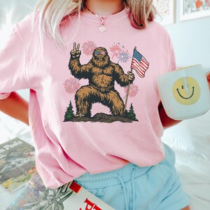 Pu&ograve; includere: Una t-shirt rosa chiaro con la grafica di un Bigfoot marrone che indossa occhiali da sole con il motivo della bandiera americana, fa il segno di vittoria e tiene in mano una bandiera americana. Il disegno include fuochi d'artificio e pini.