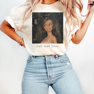 May include: A cream-colored t-shirt featuring a square graphic of a doll with long brown hair wearing a pink top. Below the graphic, the text reads, y'all need Jesus. The shirt is paired with light blue denim jeans with distressed details.